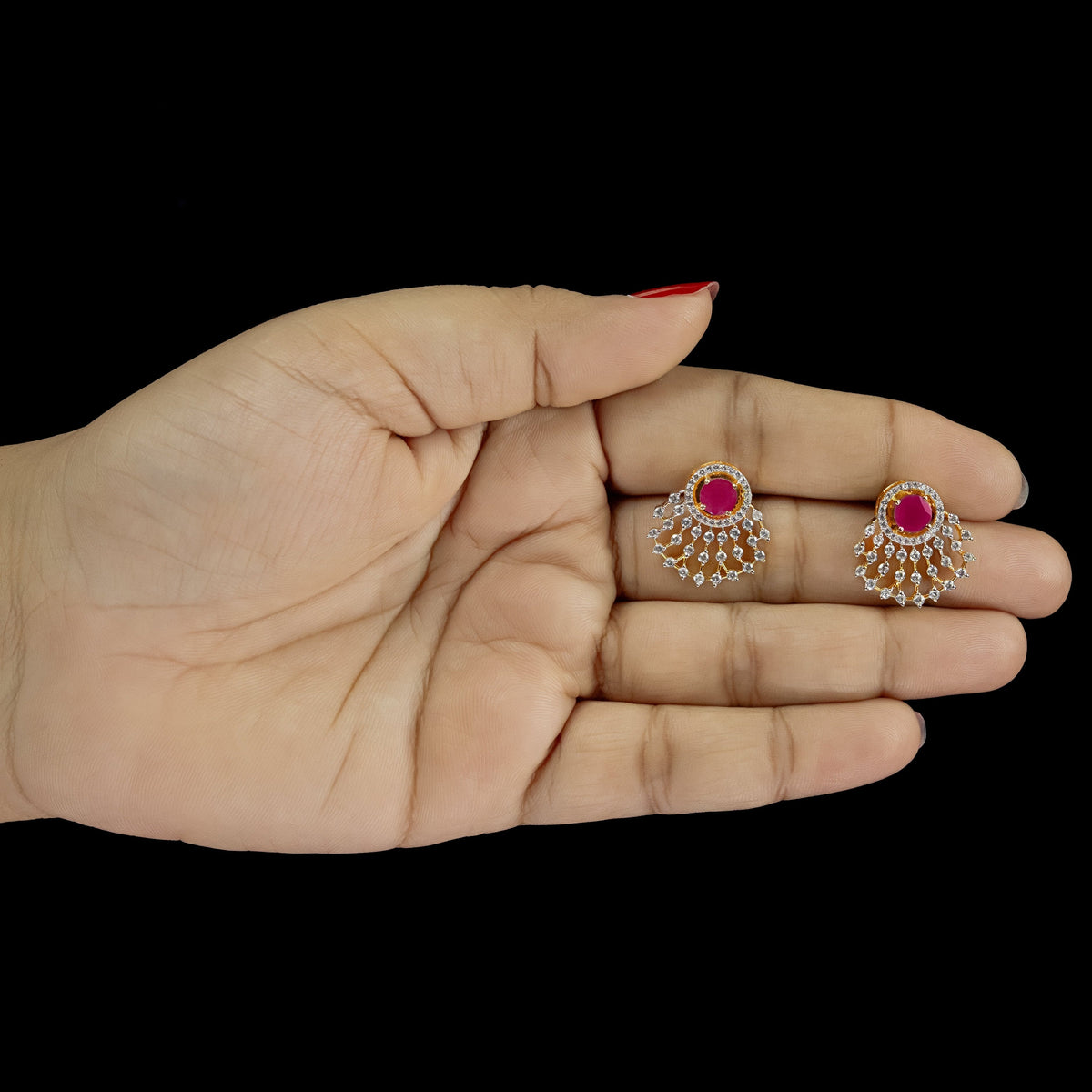 A pair of gold-toned stud earrings with central pinkish-red stones and surrounding clear stones, displayed on an open hand against a solid black background.