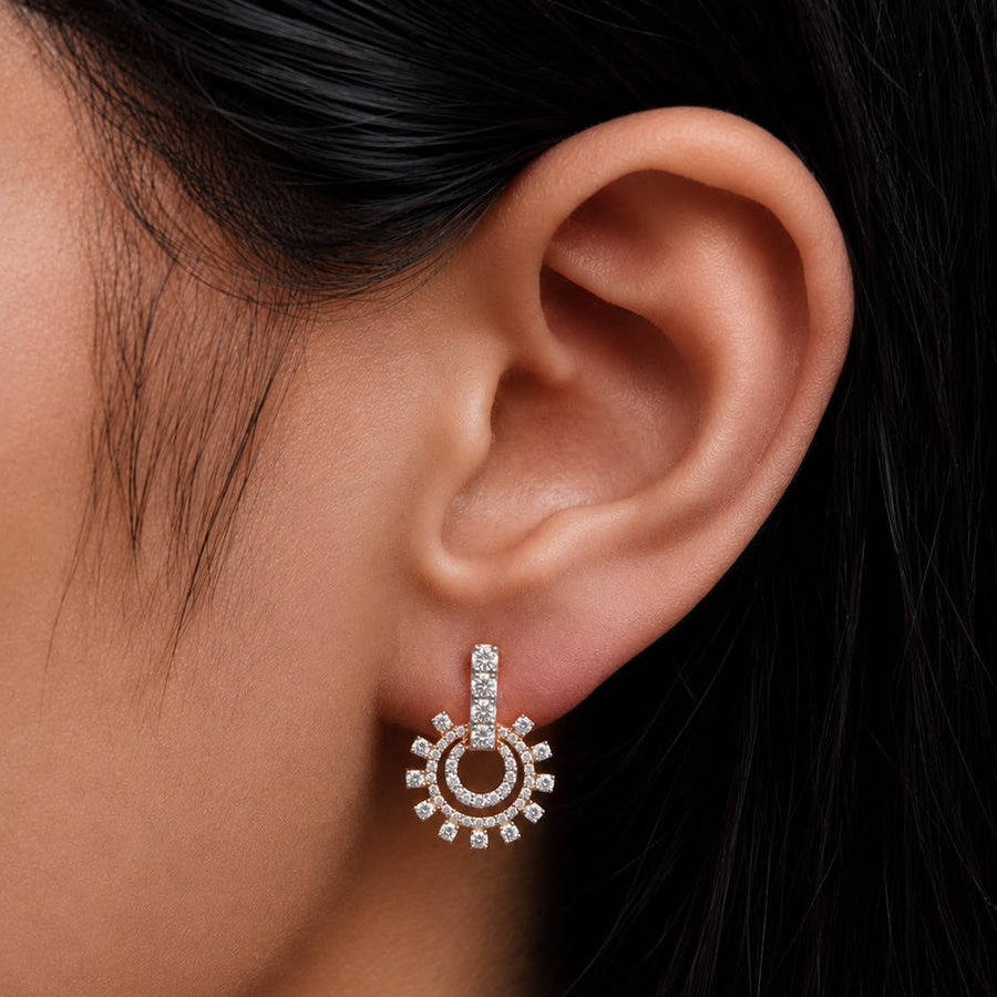 A close-up of a model's ear wearing a small gold-toned dangle earring adorned with clear stones in a sunburst design.