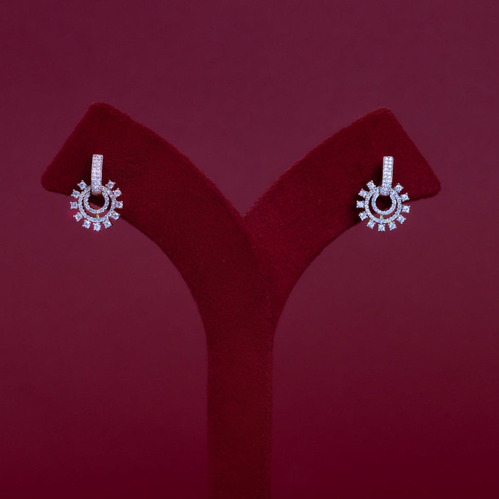 A pair of silver-tone drop earrings with circular sunburst designs set with clear stones, displayed on a red velvet jewelry stand against a dark red background.