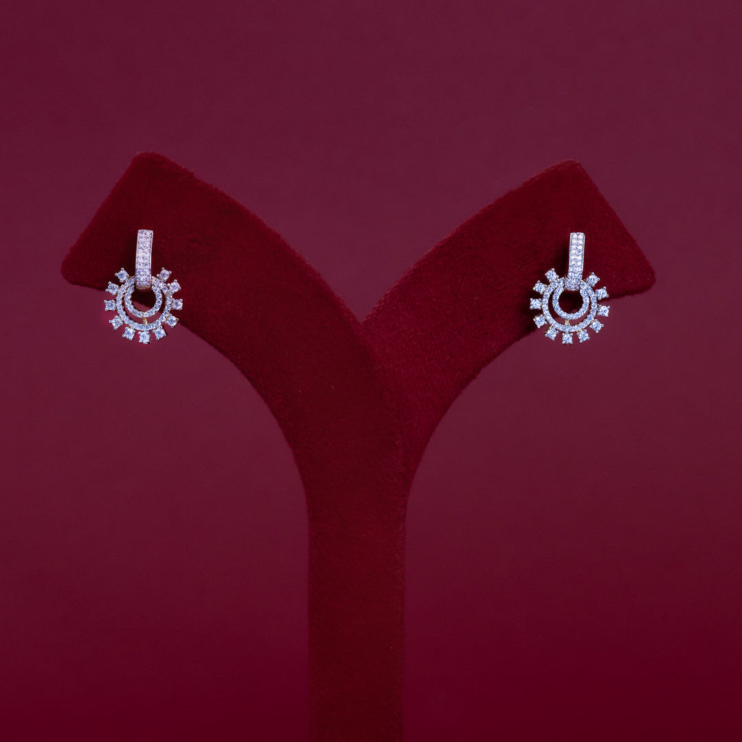 A pair of silver-tone drop earrings with circular sunburst designs set with clear stones, displayed on a red velvet jewelry stand against a dark red background.